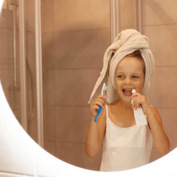 Indoor shot of little girl brushing teeth in bathroom, looking at her reflection in the mirror with excited facial expression, , wearing white t shirt and wrapped her hair in towel.